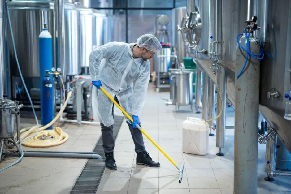 Profissional de limpeza técnica com uniforme branco e touca higiênica esfregando o piso de uma fábrica cercada por tanques de aço inoxidável.